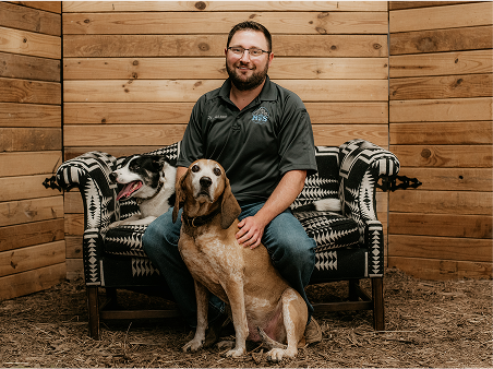 A man seated on a chair, accompanied by two dogs, enjoying a moment of companionship in a serene setting.