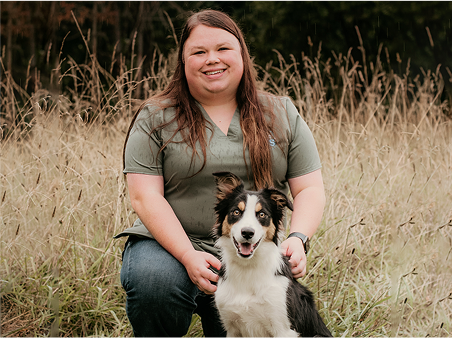 A woman kneels in tall grass, affectionately interacting with her dog beside her.