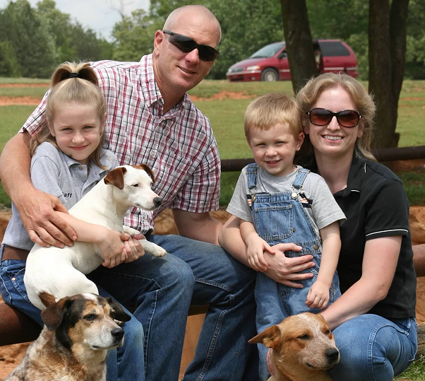 A man, woman, and two children smile while posing with their two dogs in a sunny outdoor setting.