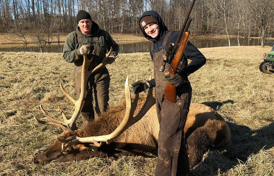 Two men stand in a field, smiling beside a large elk they have harvested.