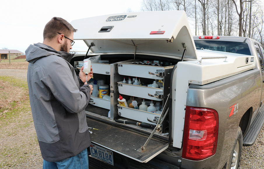 A man stands in the back of a truck, looking out over the surroundings with a focused expression.