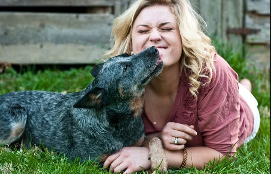 A woman lies on the grass, smiling, while her dog rests beside her, enjoying a sunny day outdoors.